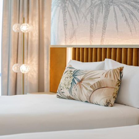 Amora Hotel Брісбен Екстер'єр фото The photo shows a stylish hotel room featuring two neatly made beds with white linens. Each bed has several pillows, one of which has a decorative cover with a tropical plant pattern. Behind the beds is a modern headboard, and the wall is decorated w