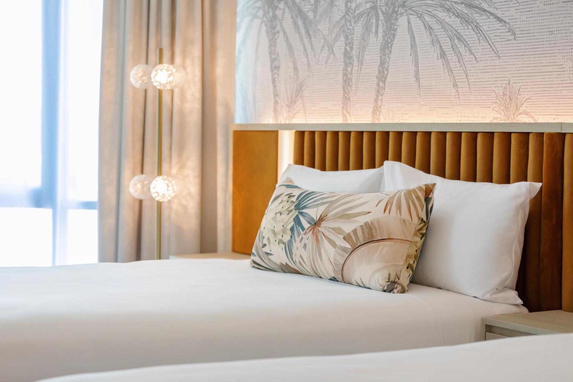 Amora Hotel Брісбен Екстер'єр фото The photo shows a stylish hotel room featuring two neatly made beds with white linens. Each bed has several pillows, one of which has a decorative cover with a tropical plant pattern. Behind the beds is a modern headboard, and the wall is decorated w