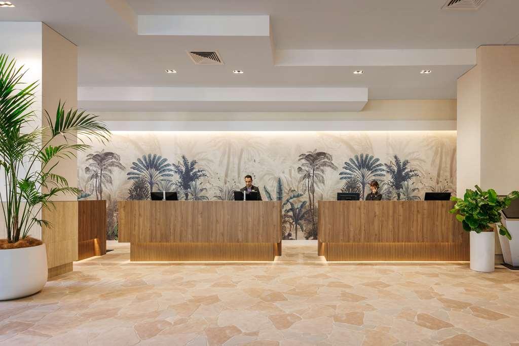 Amora Hotel Брісбен Інтер'єр фото The photo shows a modern reception area with two wooden reception desks. Behind the desks, there is a decorative wall featuring a large mural of tropical plants and palm trees, creating a serene atmosphere. The space is well-lit with overhead lights,